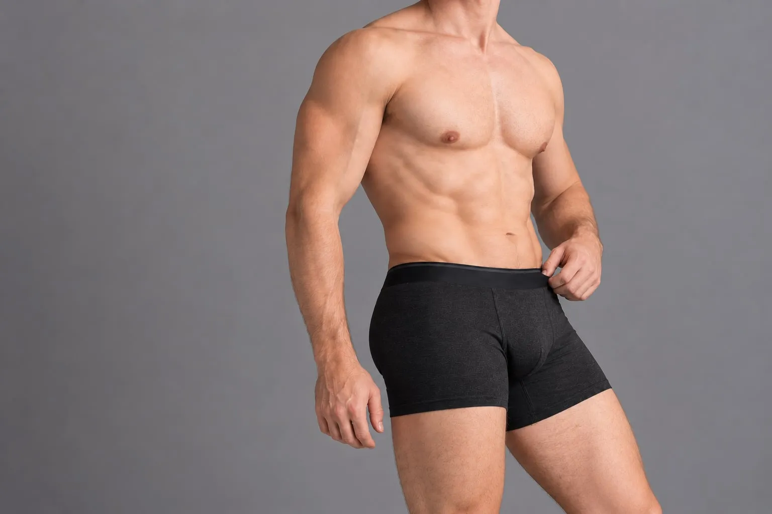 Homem de cueca boxer curta preta, de pé, ajustando o cós, em estúdio de fundo cinza